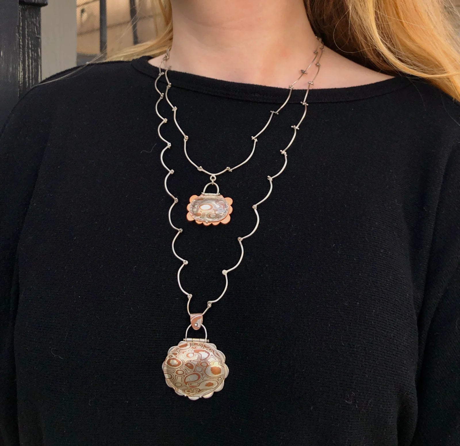 Person wearing a black top with a long, ornate necklace featuring two pendants.
